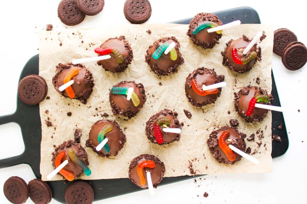 Twelve Worms and Dirt Cake Pops&mdash;chocolate-covered apples with cookie crumbs and gummy worms on sticks&mdash;are arranged on parchment paper, with sandwich cookies scattered around.