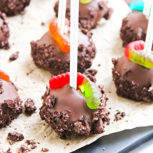 Worms and Dirt Cake Pops on sticks, coated in crushed cookies and topped with gummy worms, are arranged on parchment paper.