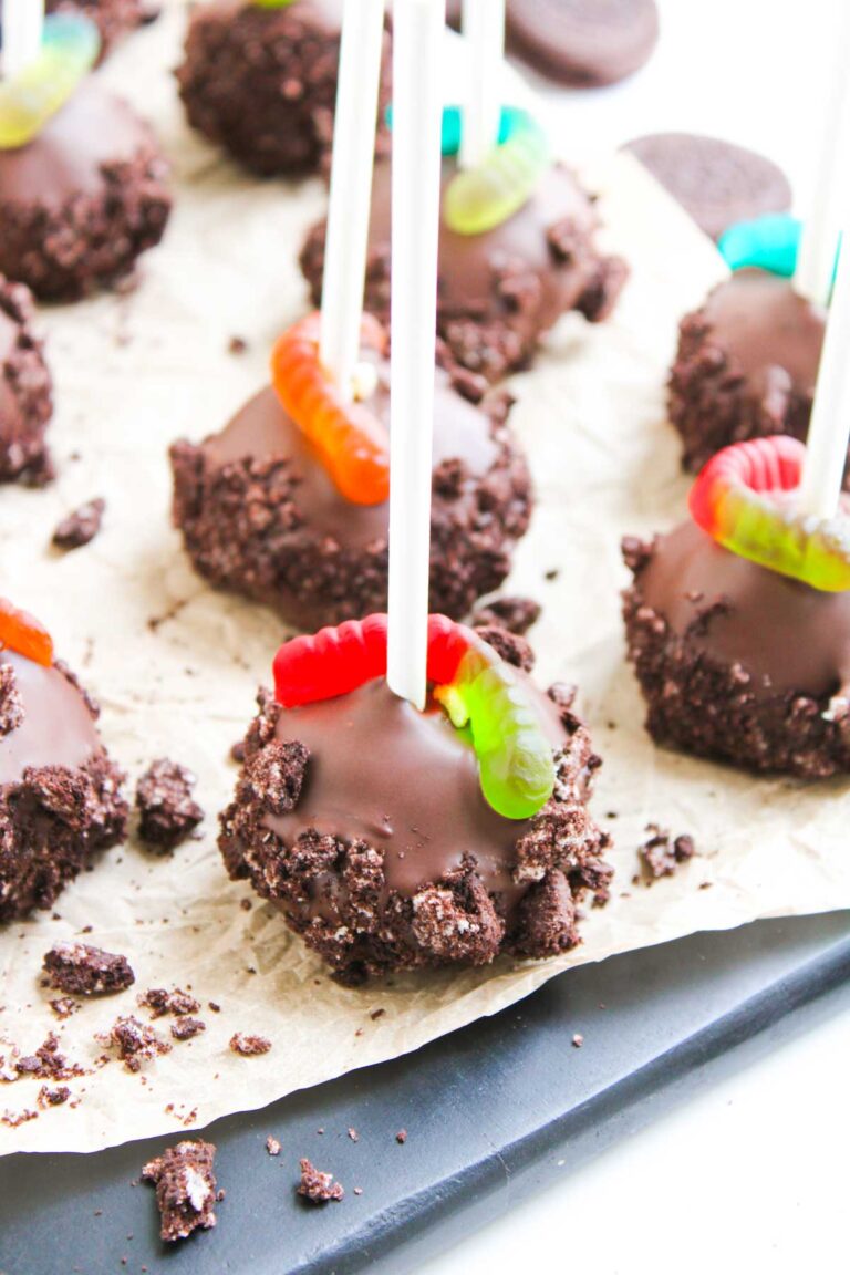 Worms and Dirt Cake Pops on sticks, coated in crushed cookies and topped with gummy worms, are arranged on parchment paper.