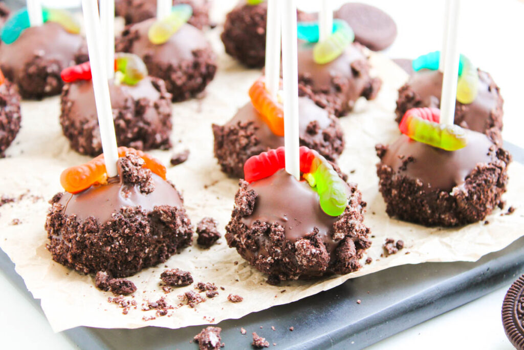 Worms and Dirt Cake Pops feature chocolate cake pops with Oreo crumbs on sticks, topped with gummy worms and arranged on parchment paper.