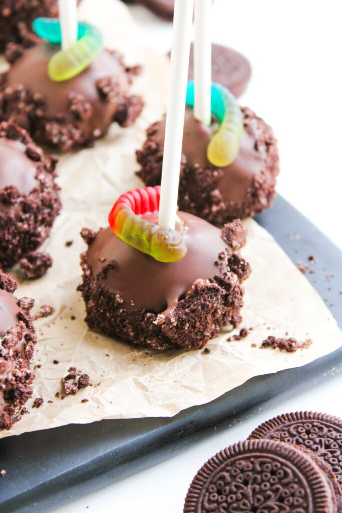 Worms and Dirt Cake Pops: Chocolate-coated cake pops on sticks, rolled in crushed cookies and topped with gummy worms, displayed on parchment paper with Oreo cookies nearby.