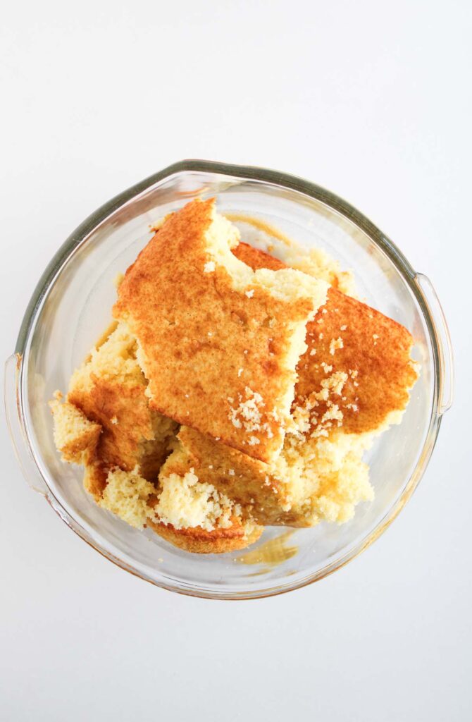A glass bowl containing several pieces of crumbled yellow cornbread on a white background, similar in texture to the base of Worms and Dirt Cake Pops.
