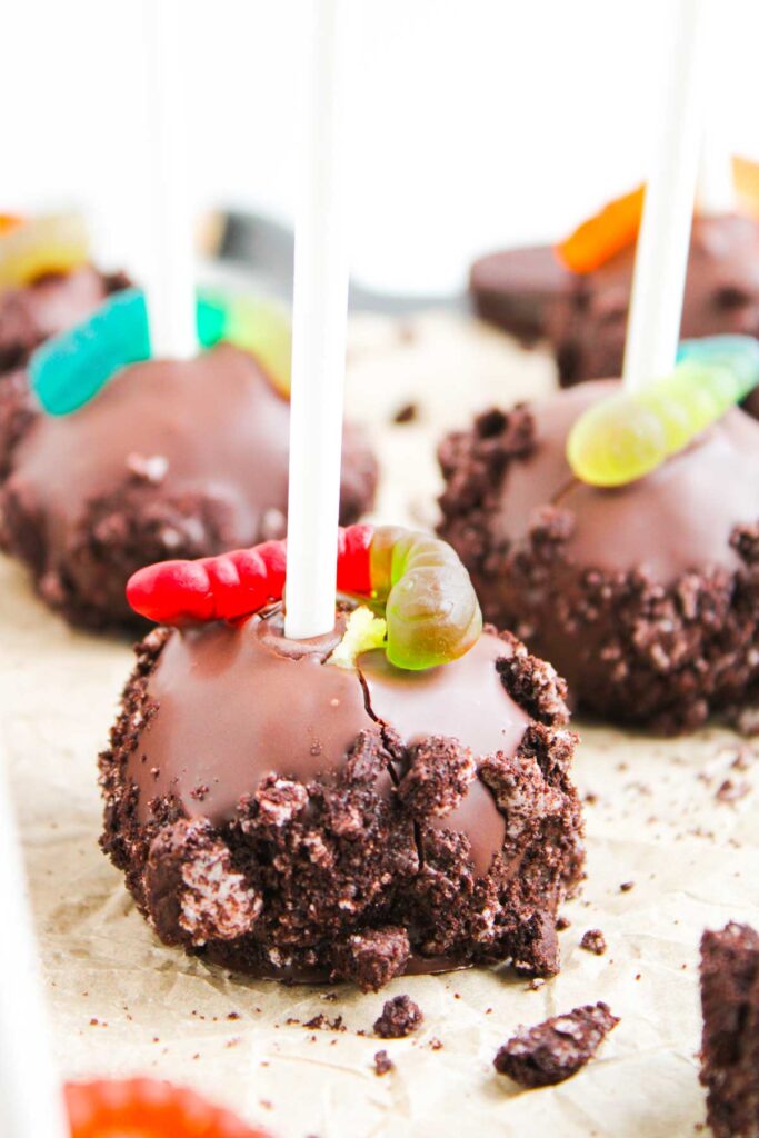 Worms and Dirt Cake Pops&mdash;chocolate-covered cake pops rolled in crushed cookies and topped with gummy worms, artfully displayed on parchment paper.
