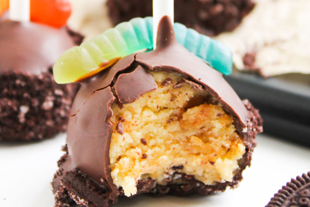 A chocolate-covered Worms and Dirt Cake Pop with a bite taken out, topped with a gummy worm, is displayed on a white surface.