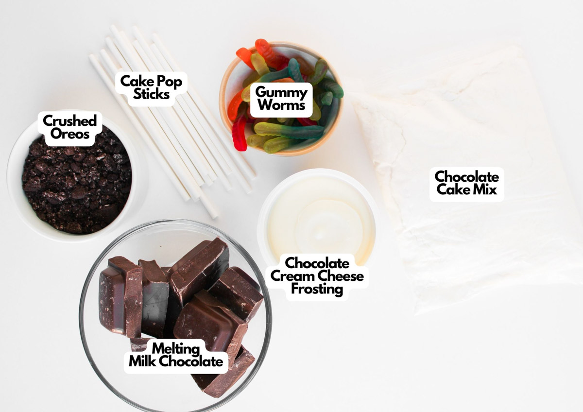 Ingredients for Worms and Dirt Cake Pops are arranged on a white surface: crushed Oreos, cake pop sticks, gummy worms, chocolate cake mix, chocolate cream cheese frosting, and melting milk chocolate.