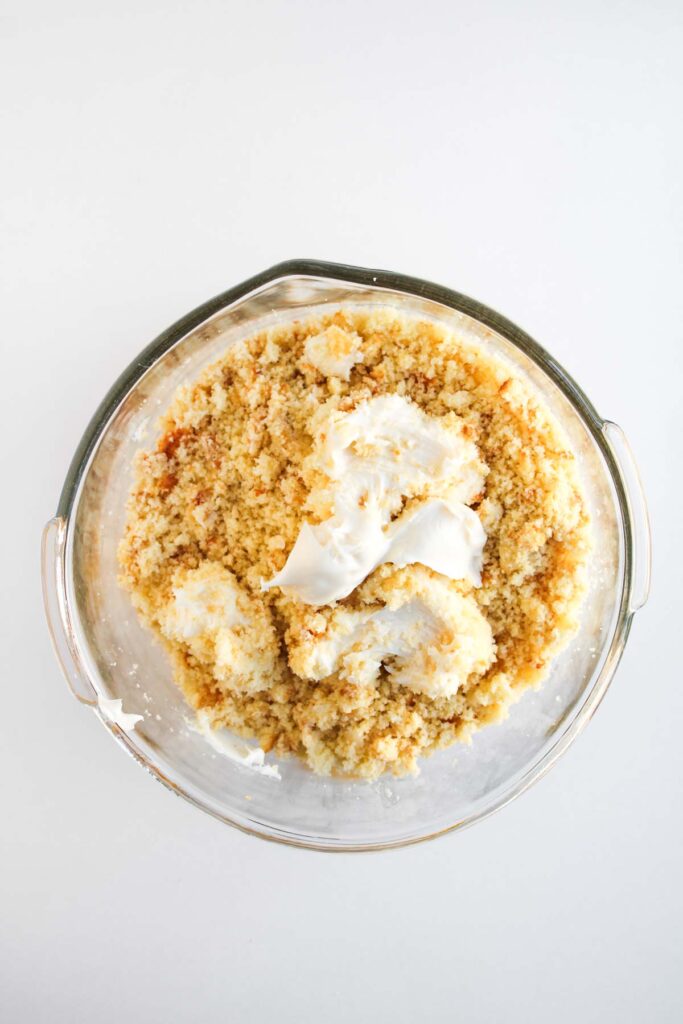A glass bowl filled with crumbled cake and dollops of white frosting on a white surface, resembling the sweet layers found in Worms and Dirt Cake Pops.