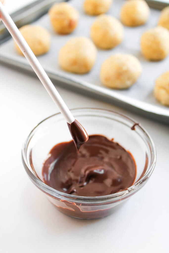 A bowl of melted chocolate with a spoon above it, ready for dipping Worms and Dirt Cake Pops; a tray of uncoated dough balls sits in the background.