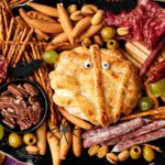 A Halloween-themed charcuterie board featuring a Camembert Cheese Mummy with candy eyes, surrounded by meats, breadsticks, grapes, nuts, olives, and a plastic spider.