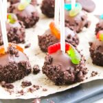 Worms and Dirt Cake Pops coated in chocolate and cookie crumbs, topped with gummy worms, are arranged on parchment paper with extra crumbs scattered around for a fun and playful treat.