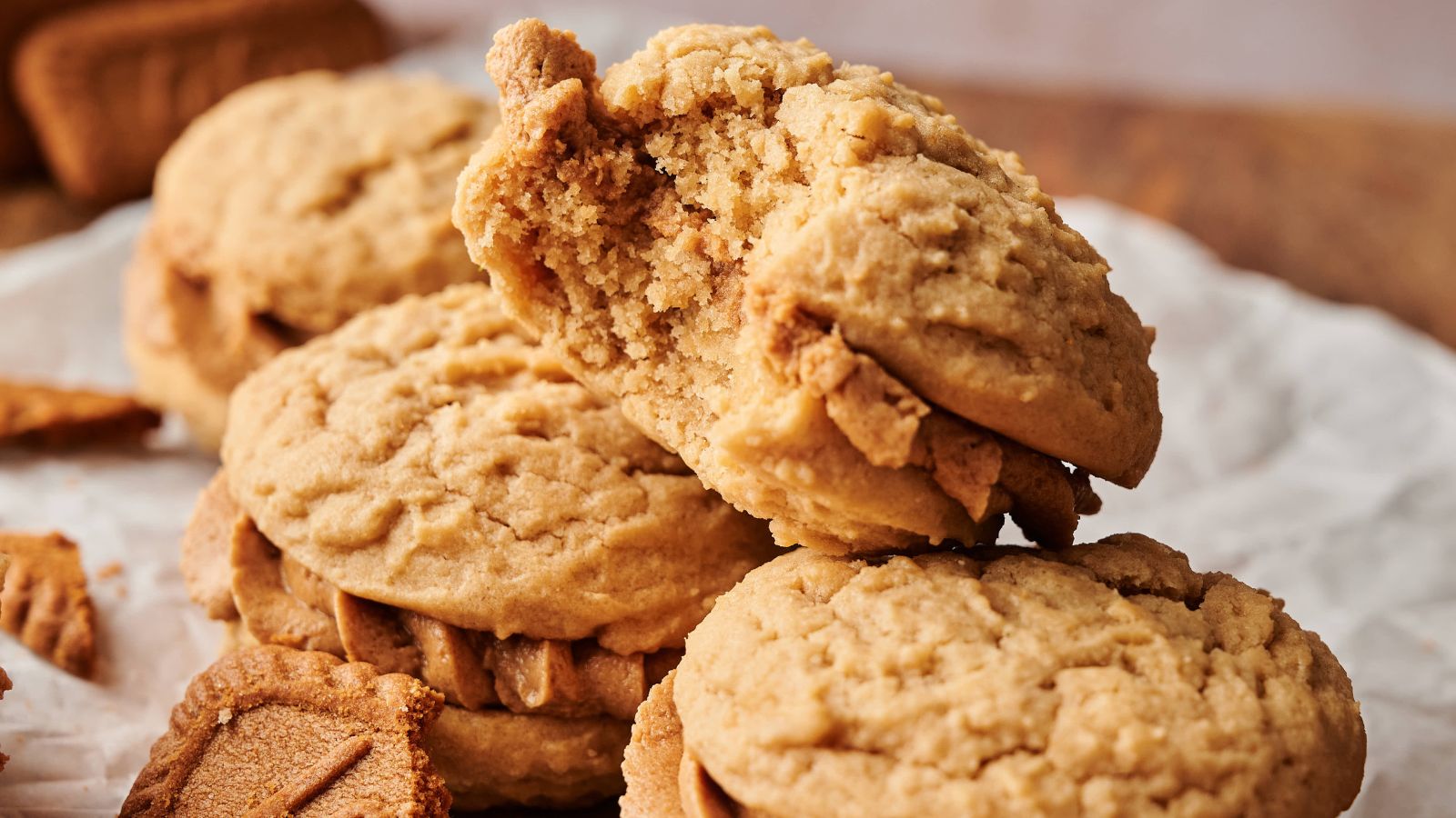 Three peanut butter sandwich cookies are stacked, with the top cookie showing a large bite taken out. Crumbs and cookie pieces are scattered on a parchment-lined surface.