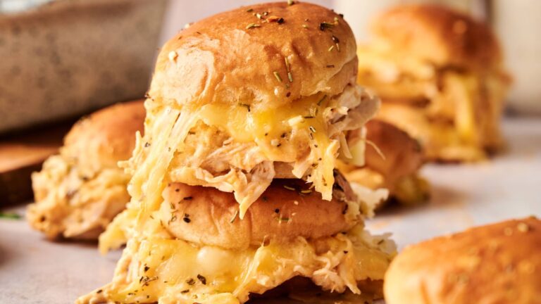 A close-up of stacked sliders filled with shredded chicken and melted cheese, topped with herbs, on a light surface.