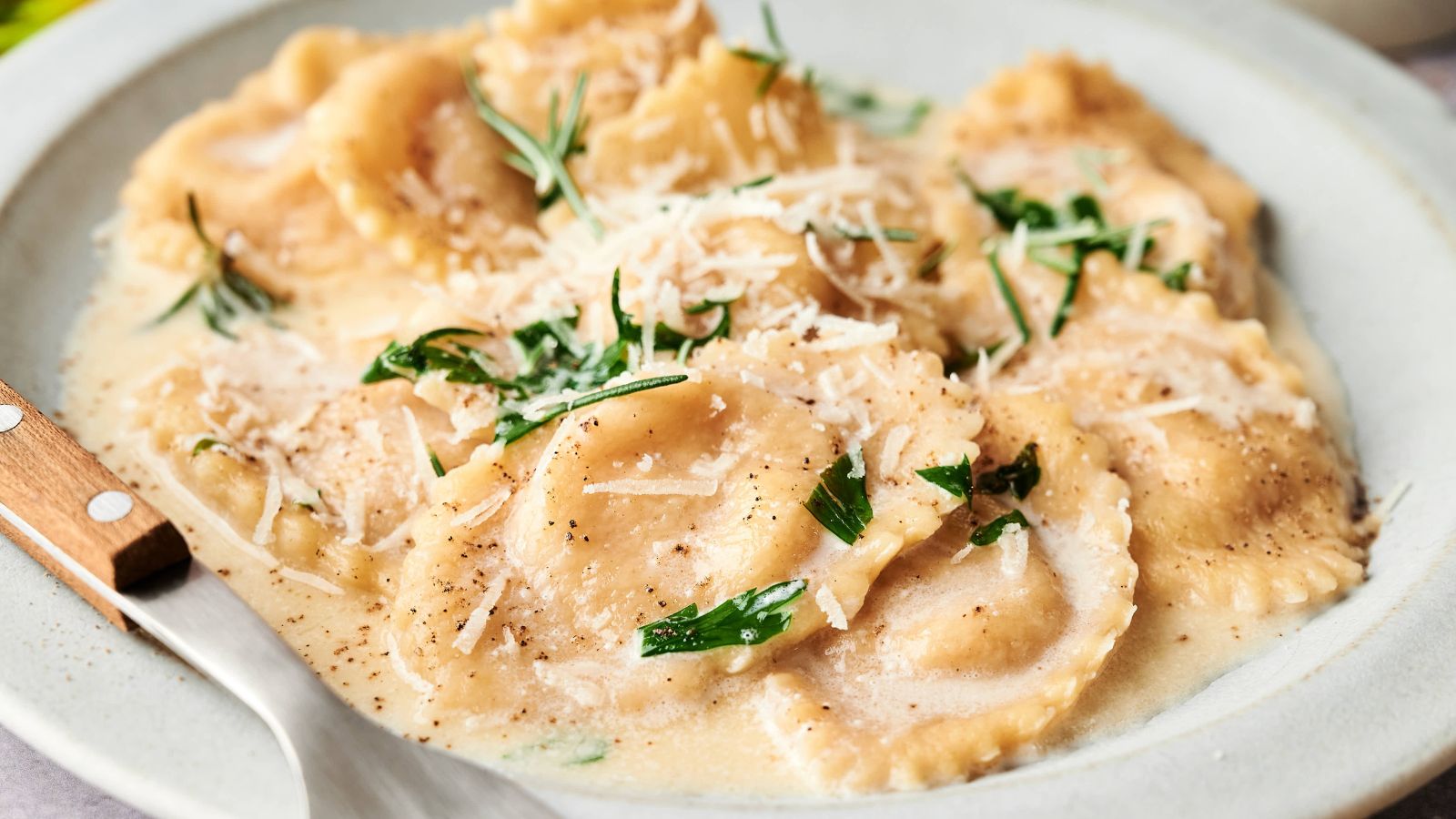 A plate of ravioli topped with creamy sauce, grated cheese, and fresh herbs, served with a fork.