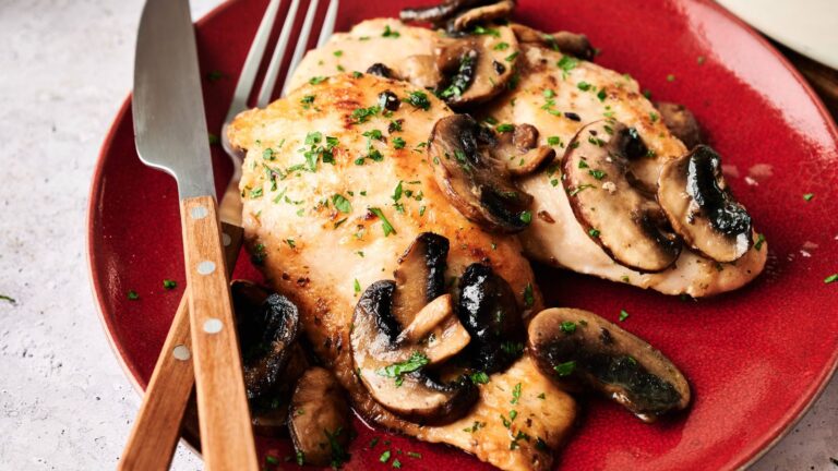 Two cooked chicken breasts topped with sautéed mushrooms and herbs are served on a red plate with a fork and knife.