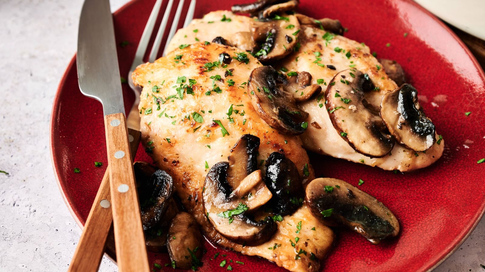 Two pieces of cooked chicken breast topped with saut&eacute;ed mushrooms and chopped herbs on a red plate, with a fork and knife beside them.