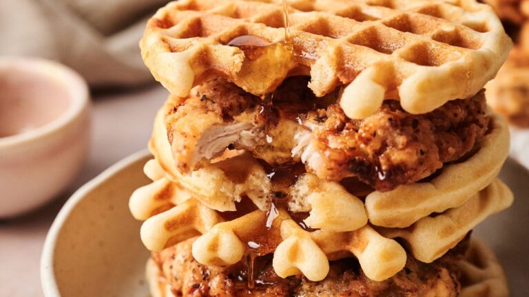 A stack of waffles and fried chicken pieces on a plate, with syrup being drizzled over the top.