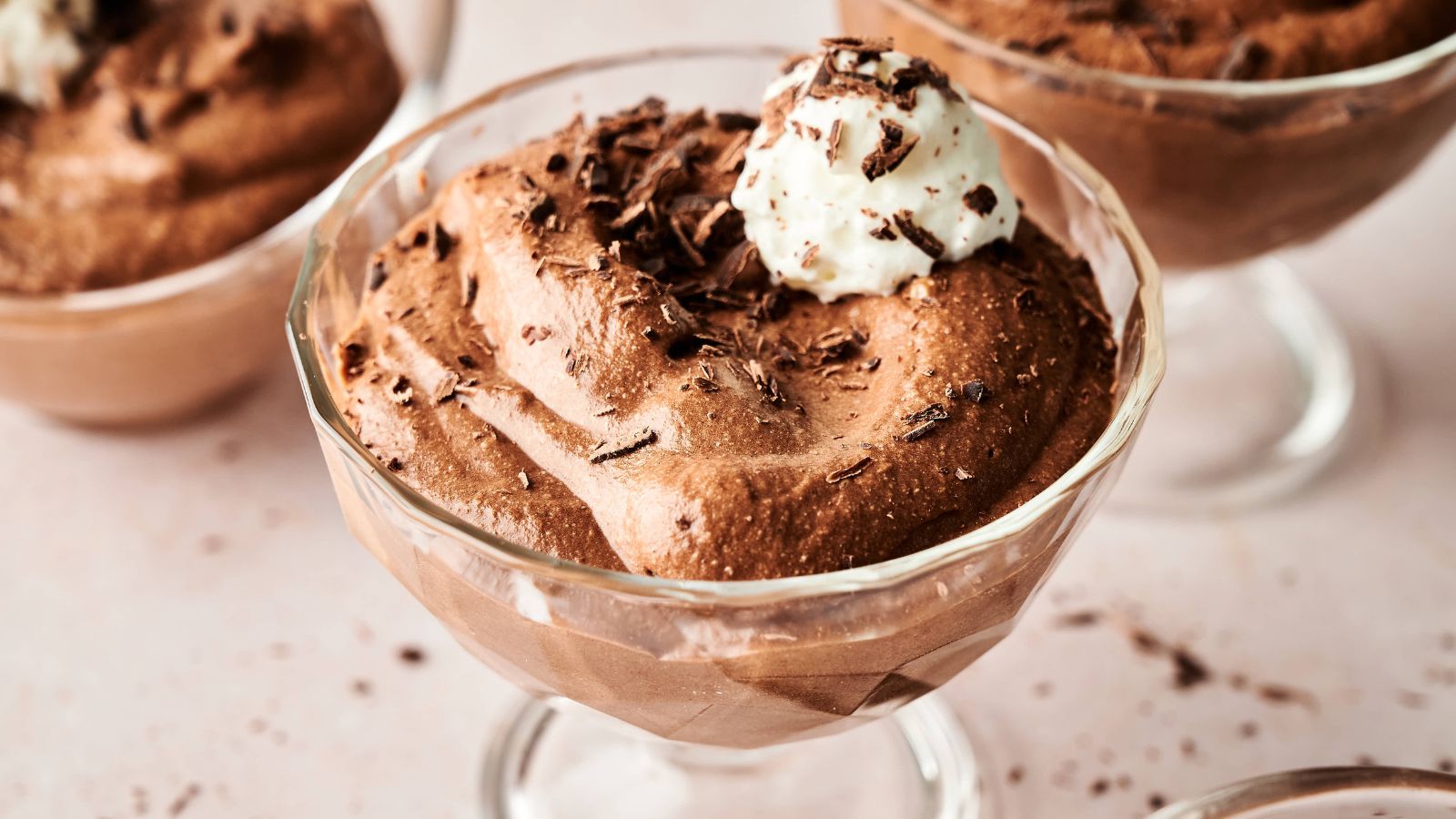 A glass dessert cup filled with chocolate mousse, topped with whipped cream and chocolate shavings, placed on a light-colored surface.