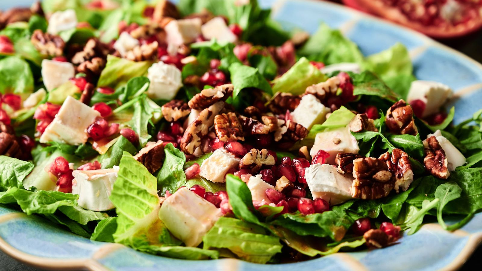 A close-up of a salad with chopped romaine lettuce, diced white cheese, pecans, and pomegranate seeds on a blue plate.