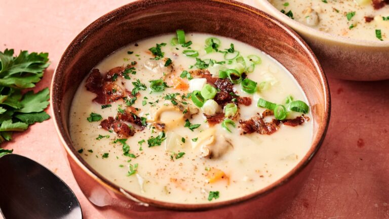 A bowl of creamy soup garnished with chopped green onions, parsley, and pieces of bacon, with a spoon and herbs nearby on a pink surface.