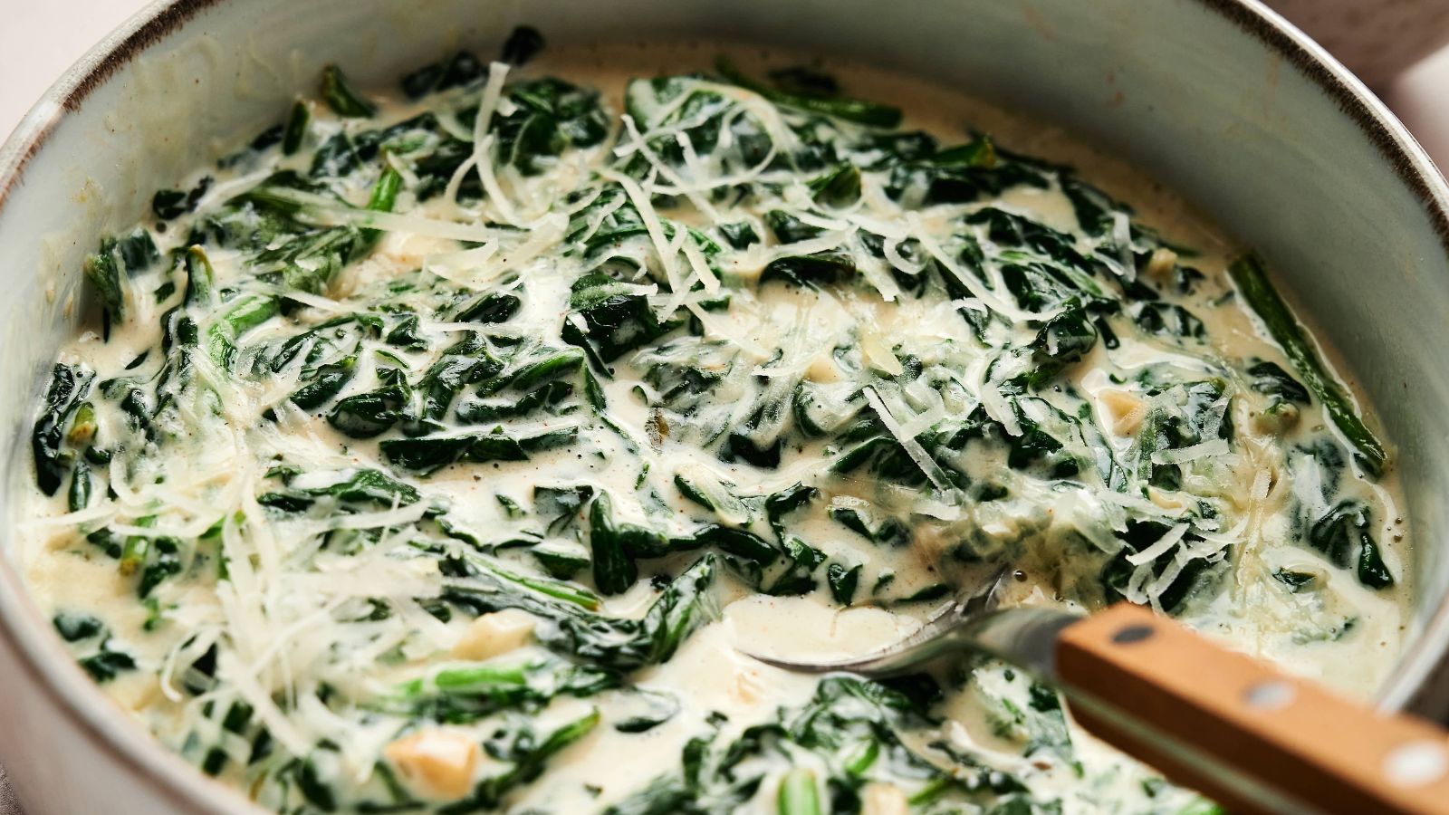 A bowl of creamy spinach topped with grated cheese, being stirred with a spoon.