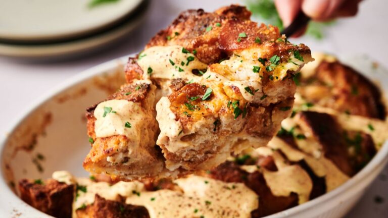A serving of bread pudding topped with creamy sauce and chopped herbs is being lifted from a white baking dish.