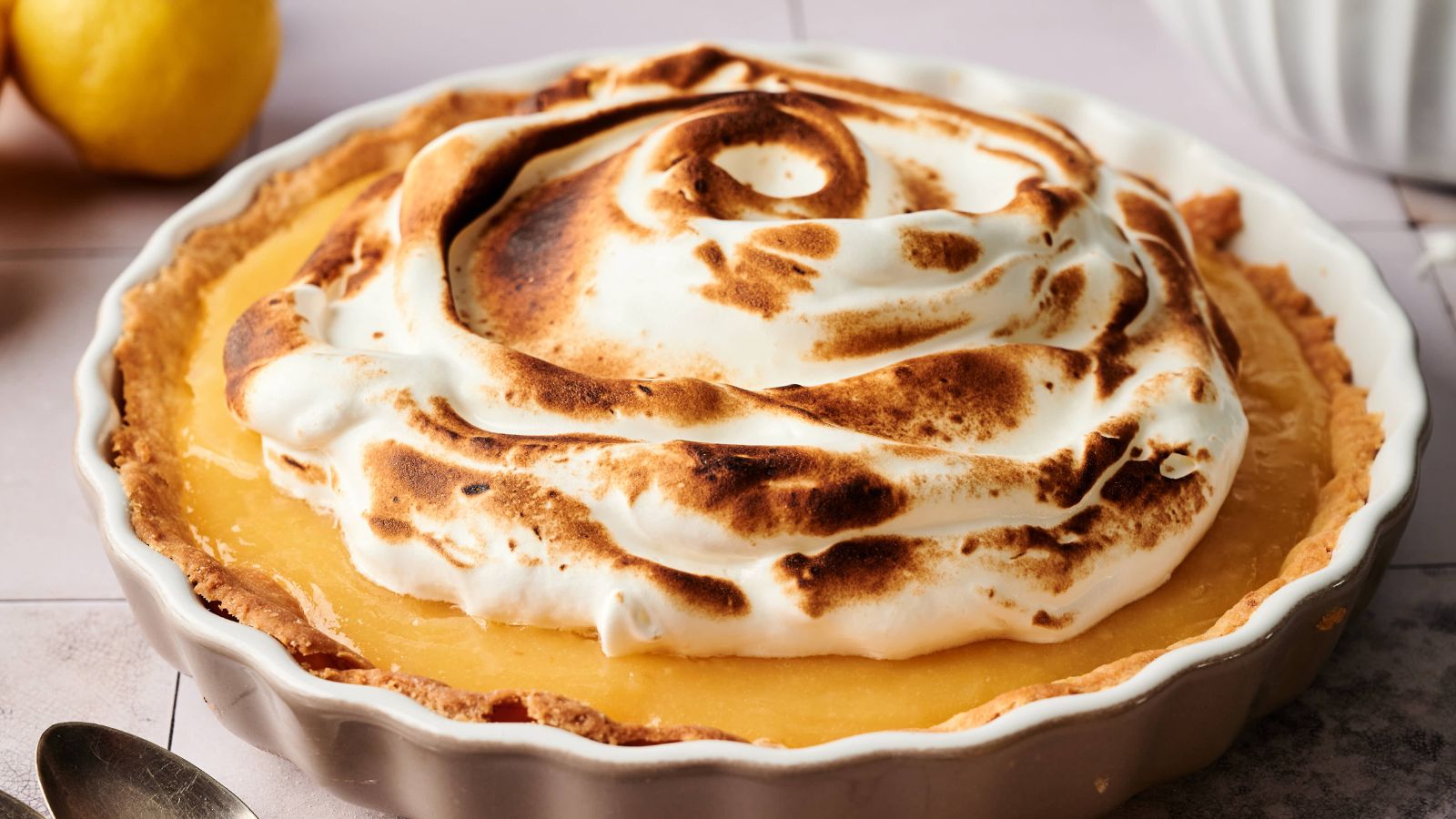 A lemon meringue pie in a white dish, topped with swirled, browned meringue, sits on a light-colored surface next to a lemon.