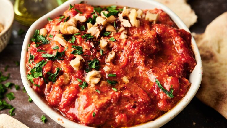A bowl of red tomato and walnut dip garnished with chopped parsley and walnuts, served with pieces of flatbread nearby.