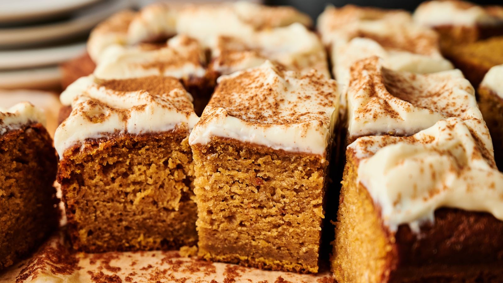 Close-up of sliced pumpkin cake topped with cream cheese frosting and a dusting of cinnamon, with plates in the background.