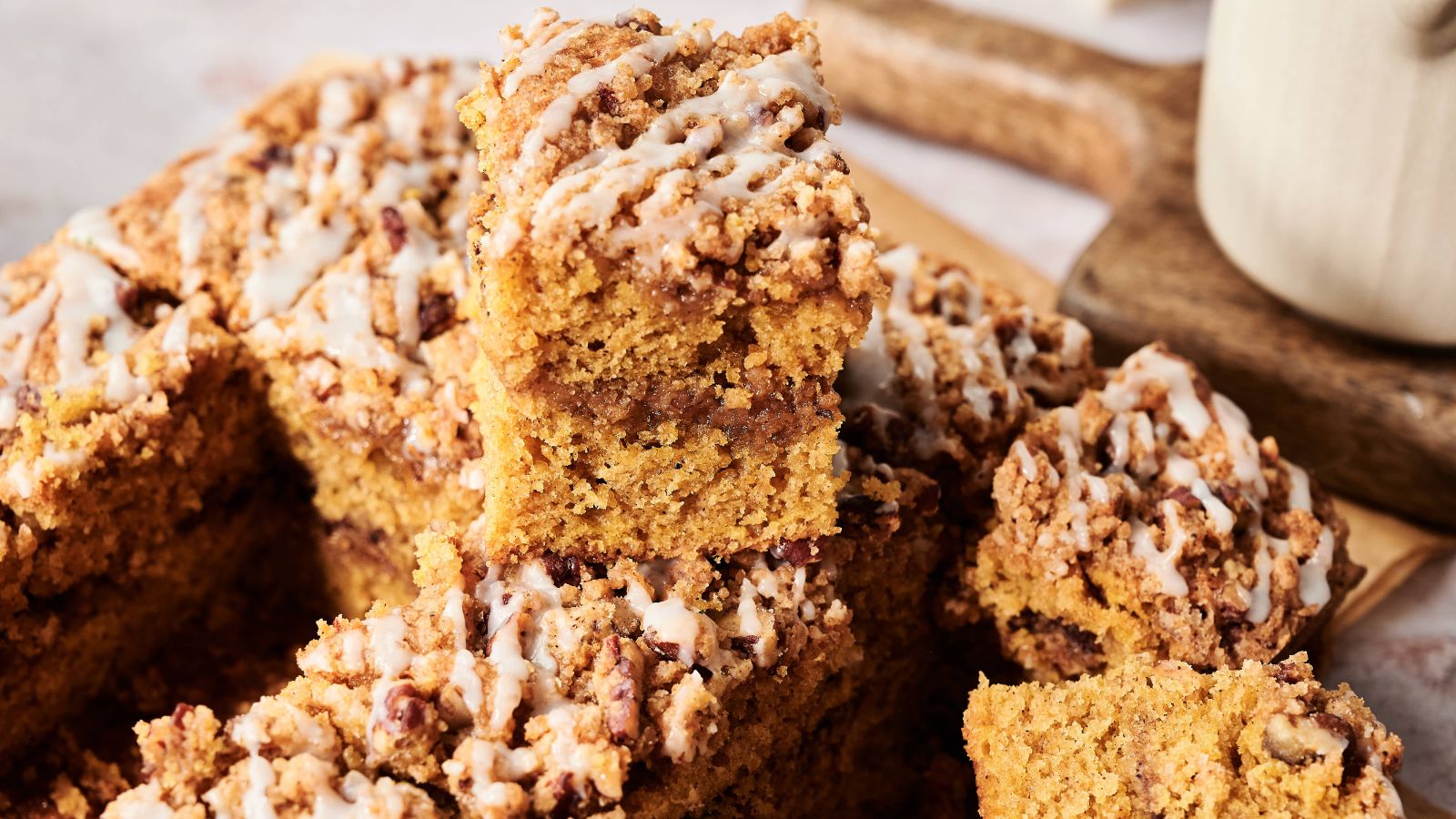 A stack of pumpkin coffee cake squares with crumb topping and white glaze, arranged on a wooden board.