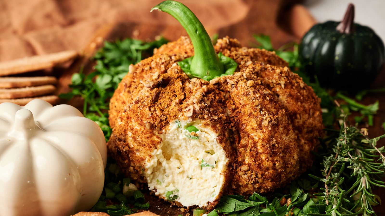 Cheese ball shaped like a pumpkin with a green stem, coated in crumbled topping, surrounded by herbs and decorative pumpkins on a wooden surface.