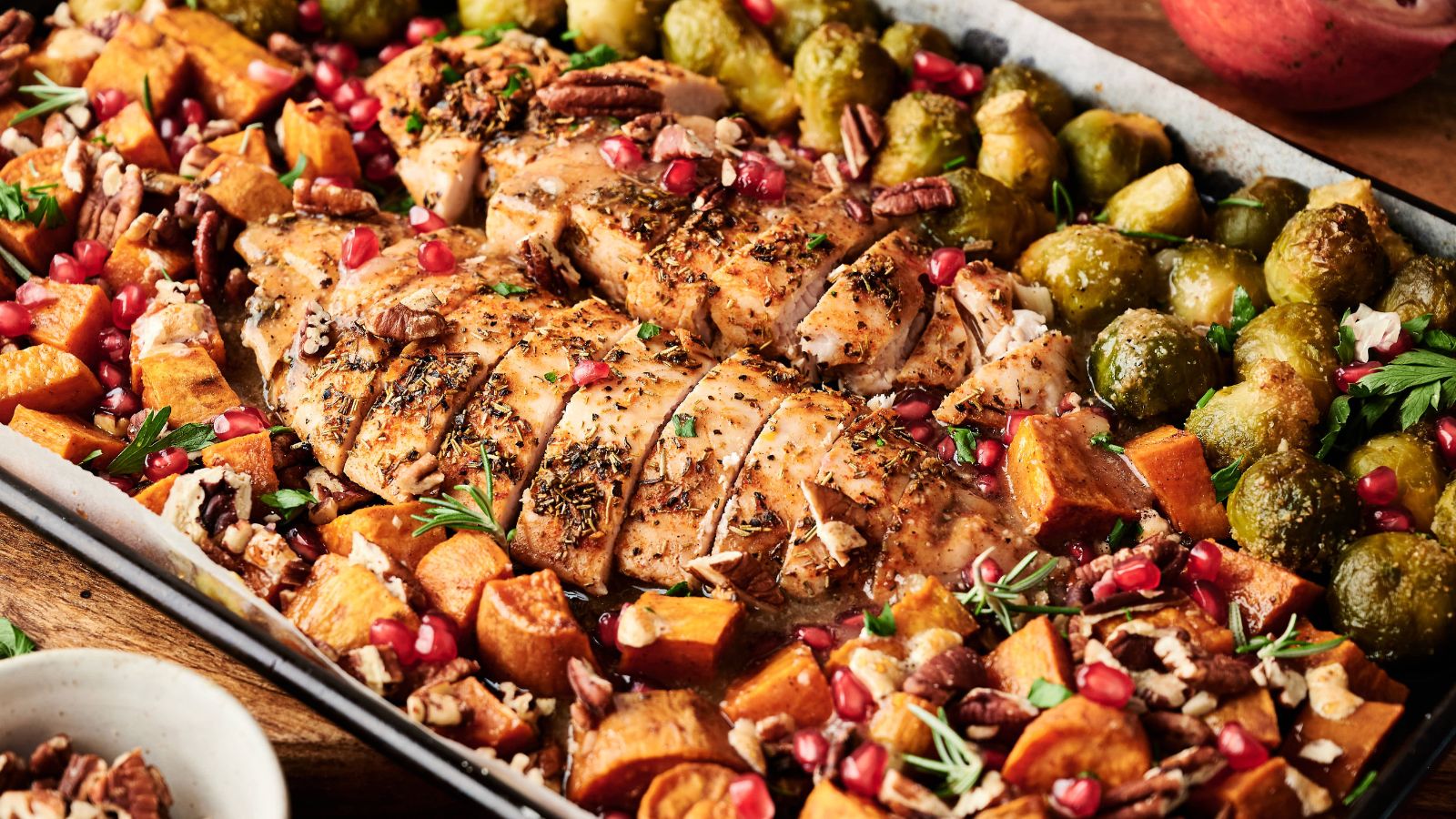 Sheet pan meal with sliced roasted chicken breast, brussels sprouts, sweet potatoes, pecans, and pomegranate seeds, garnished with herbs.