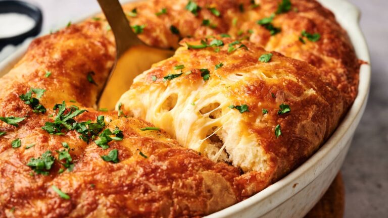 A cheesy, golden baked bread topped with chopped herbs is being served from a white oval dish with a serving utensil.