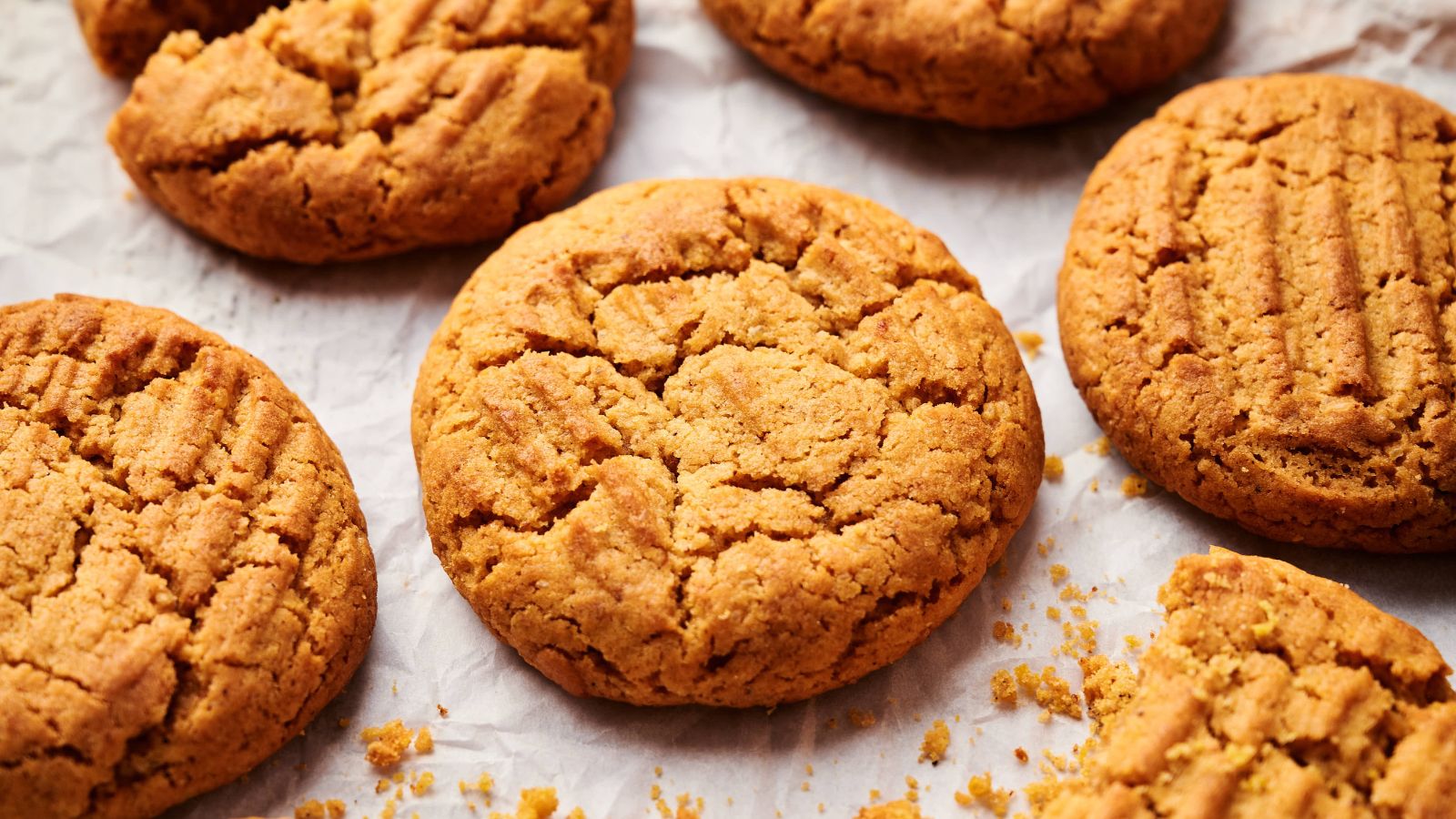 Several peanut butter cookies with a crisscross pattern on top are arranged on crumpled parchment paper, with some crumbs scattered around.