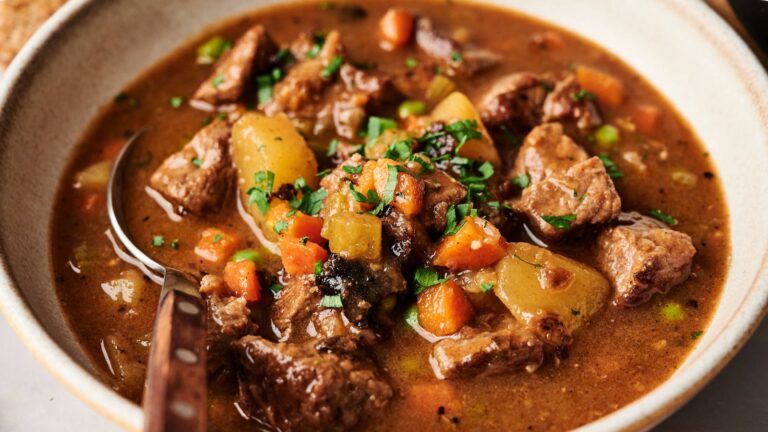 A bowl of beef stew with chunks of meat, potatoes, carrots, and peas in a brown broth, garnished with chopped parsley.