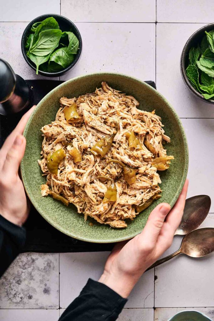 A person holds a green bowl filled with Crockpot Mississippi Chicken and green bell peppers, with bowls of fresh spinach and mint leaves nearby on a tiled surface.