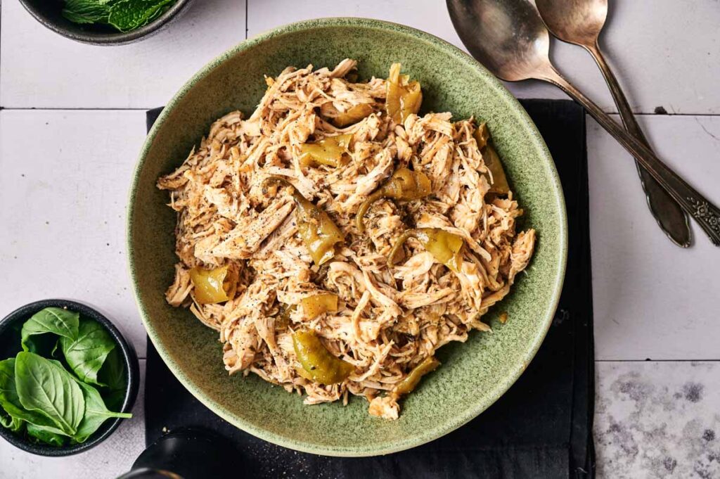 A green bowl filled with Crockpot Mississippi Chicken and sliced green peppers, placed on a black mat with utensils and a small bowl of fresh basil leaves nearby.