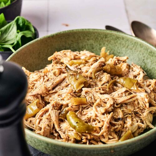 A green bowl filled with Crockpot Mississippi Chicken and sliced green bell peppers, placed on a table beside a black pepper grinder and fresh greens.