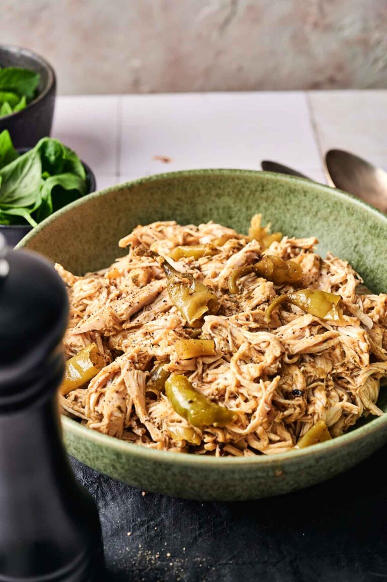 A green bowl filled with Crockpot Mississippi Chicken and sliced green bell peppers, placed on a table beside a black pepper grinder and fresh greens.