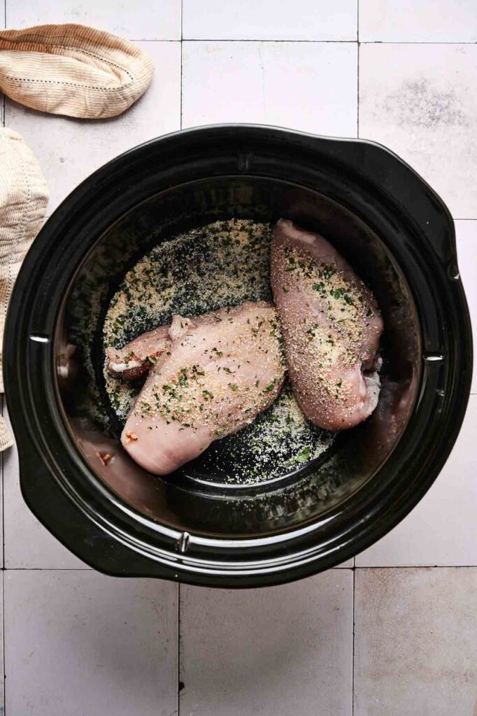 Two raw, seasoned chicken breasts in a slow cooker are ready to become delicious Crockpot Mississippi Chicken, viewed from above with a cloth napkin nearby on a tiled surface.