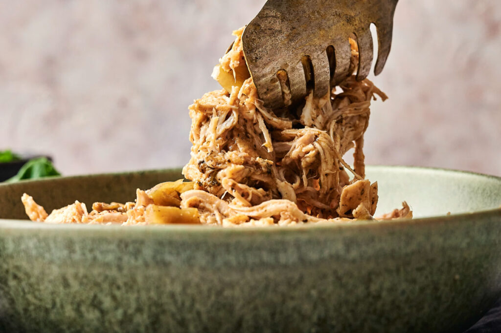 A serving fork lifts shredded Crockpot Mississippi Chicken from a green bowl.