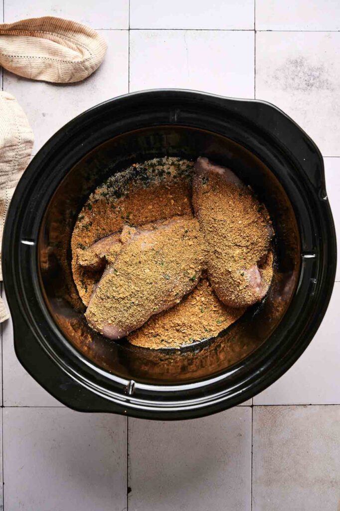 Raw, breaded chicken breasts arranged in a slow cooker, ready to become Crockpot Mississippi Chicken, with a towel visible in the corner.