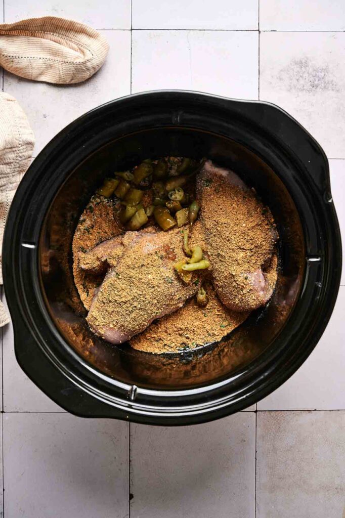 Uncooked breaded chicken breasts and chopped pickles in a black Crockpot, ready to make flavorful Crockpot Mississippi Chicken on a tiled surface.