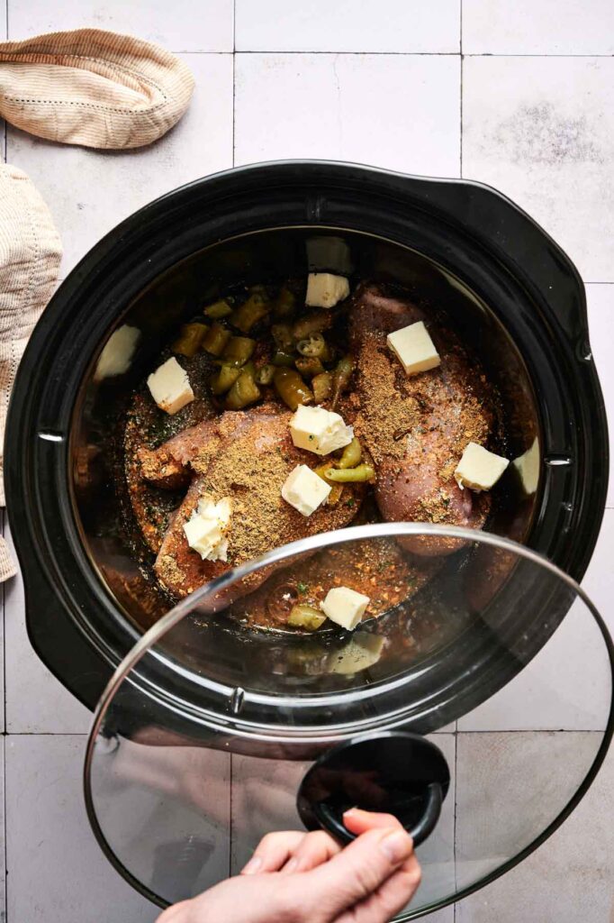 A Crockpot Mississippi Chicken dish simmers in the slow cooker with seasoned meat, cubed butter, and chopped peppers as a hand lifts the glass lid.