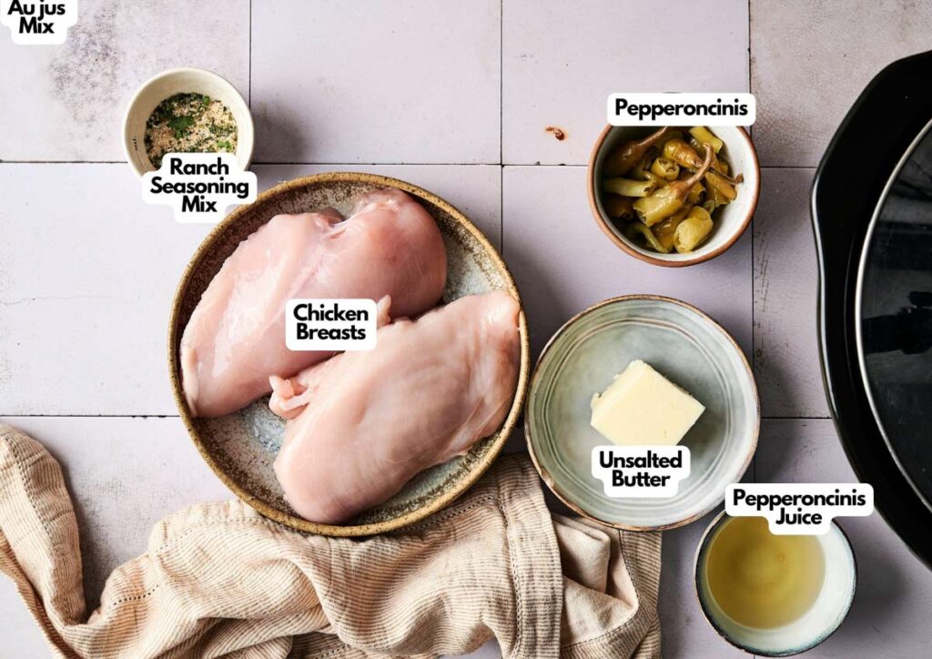Raw chicken breasts in a bowl surrounded by ranch seasoning mix, au jus mix, unsalted butter, pepperoncini peppers, and juice&mdash;perfect ingredients for making flavorful Crockpot Mississippi Chicken on your countertop.