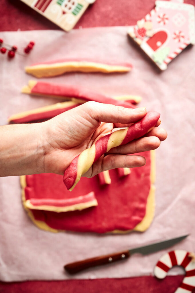 A hand holds a twisted red and yellow dough strip above a surface with rolled dough, cut strips, a knife, and holiday-themed decorations—perfect for making festive Candy Cane Cookies.