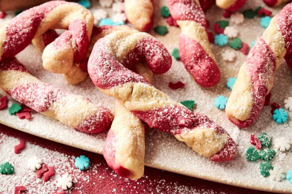 Candy Cane Cookies twisted into festive shapes are dusted with powdered sugar and artfully arranged on a plate, surrounded by colorful holiday sprinkles.