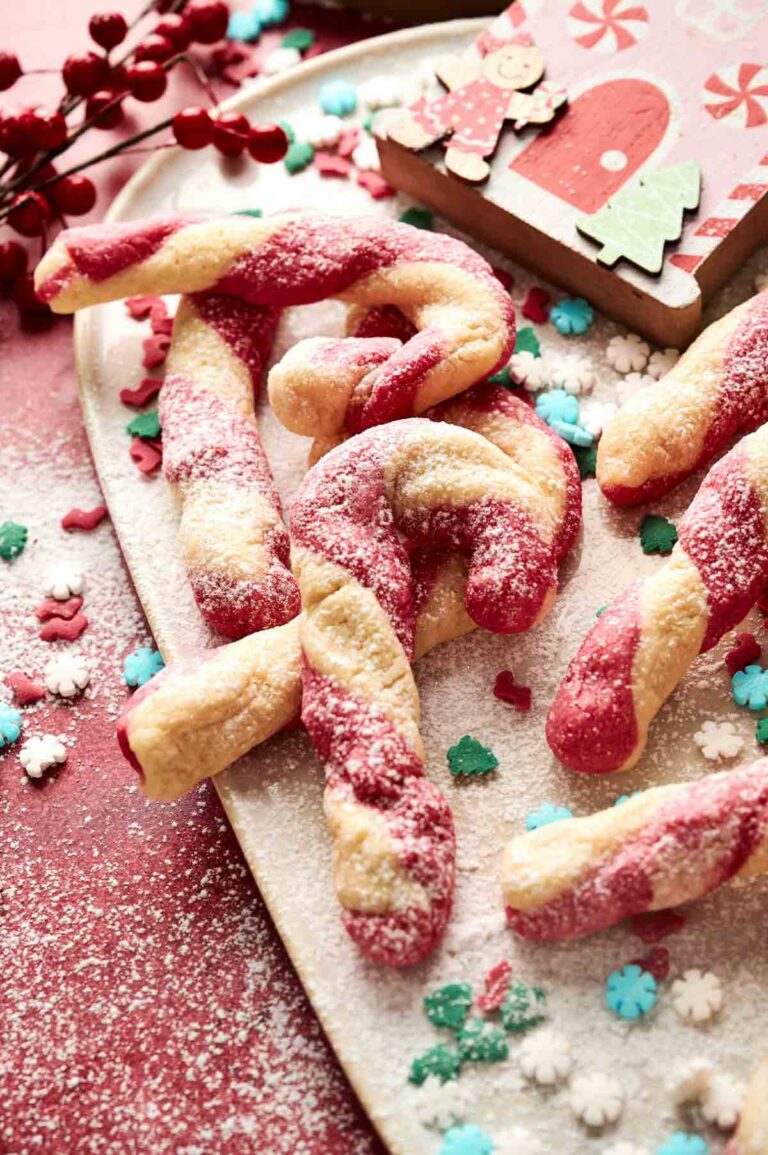 Candy Cane Cookies dusted with powdered sugar are arranged on a tray, accompanied by festive sprinkles and a holiday-themed napkin.