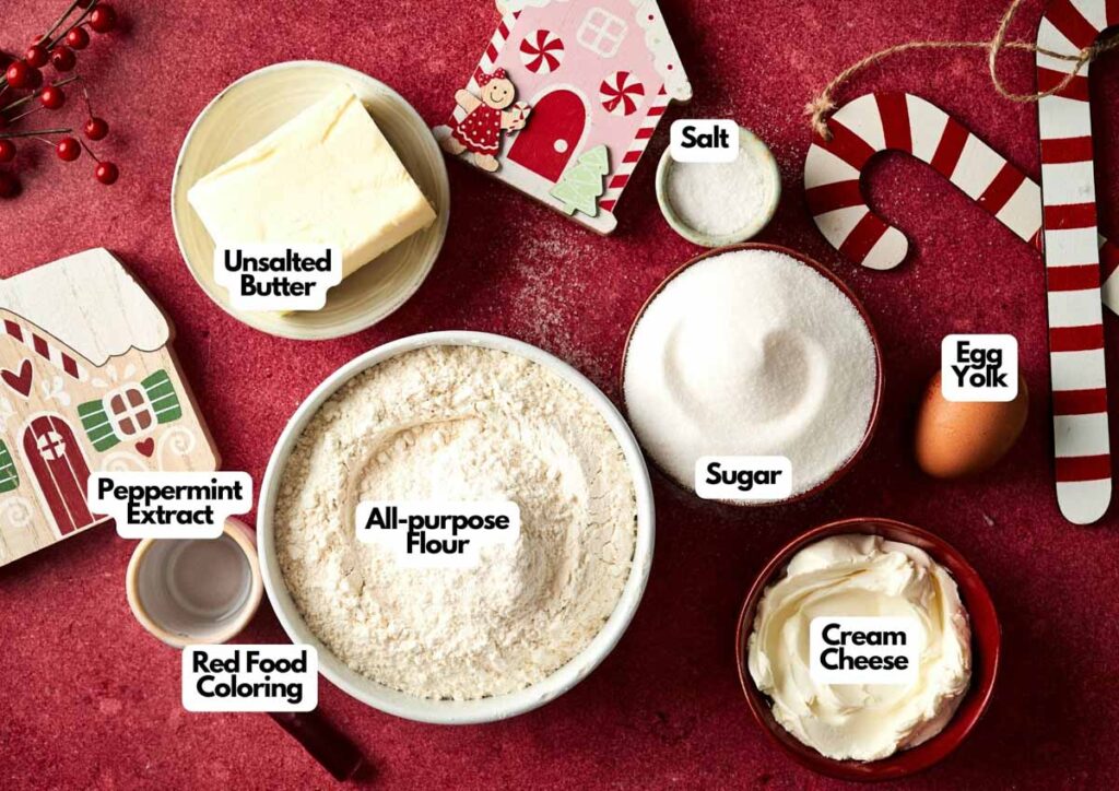 Ingredients for Candy Cane Cookies on a red surface, including unsalted butter, all-purpose flour, sugar, cream cheese, egg yolk, salt, peppermint extract, and red food coloring.