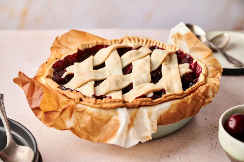 A classic Cherry Pie with a golden lattice crust sits in a dish lined with parchment paper; plates, spoons, and a fresh cherry are nearby.