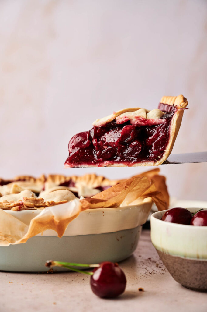 A slice of Cherry Pie is being lifted from a pie dish, its rich cherry filling oozing out, while a bowl of fresh cherries sits nearby.