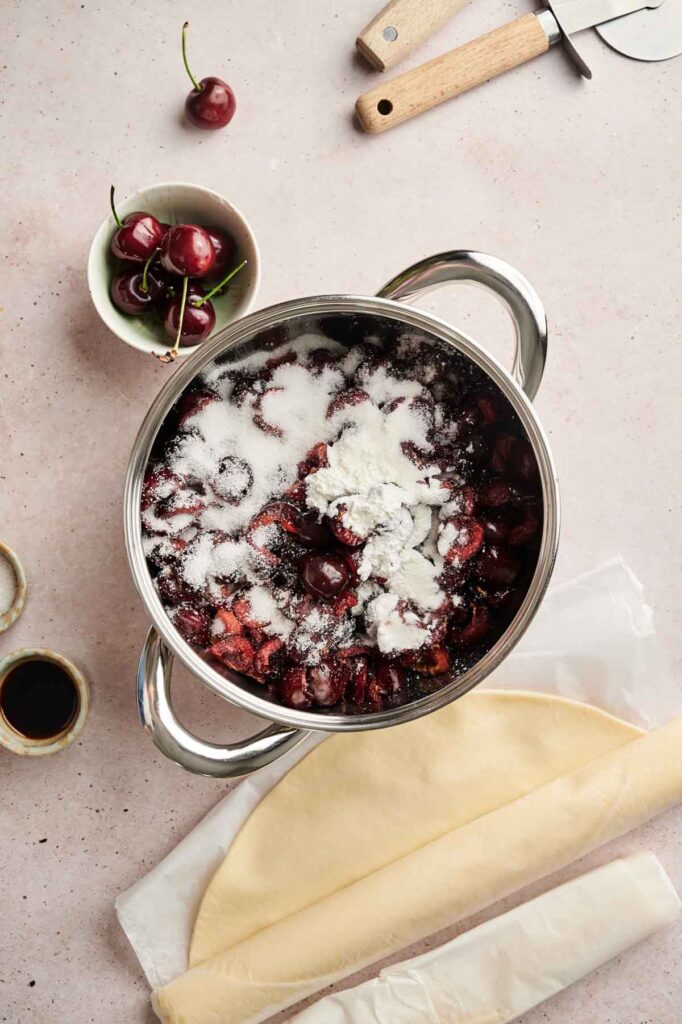 A pot filled with cherries, sugar, and flour sits on a counter next to pie crust dough, a bowl of fresh cherries, a small bowl of vanilla, and kitchen utensils&mdash;everything ready for baking the perfect Cherry Pie.